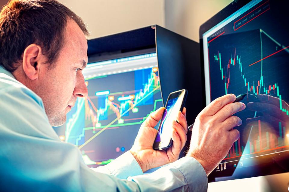 A professional money manager using a stylus and smartphone to analyze a stock chart displayed on a computer screen.