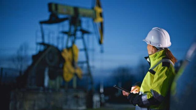 Engineer looking at machinery while using tablet PC at oil industry against sky
